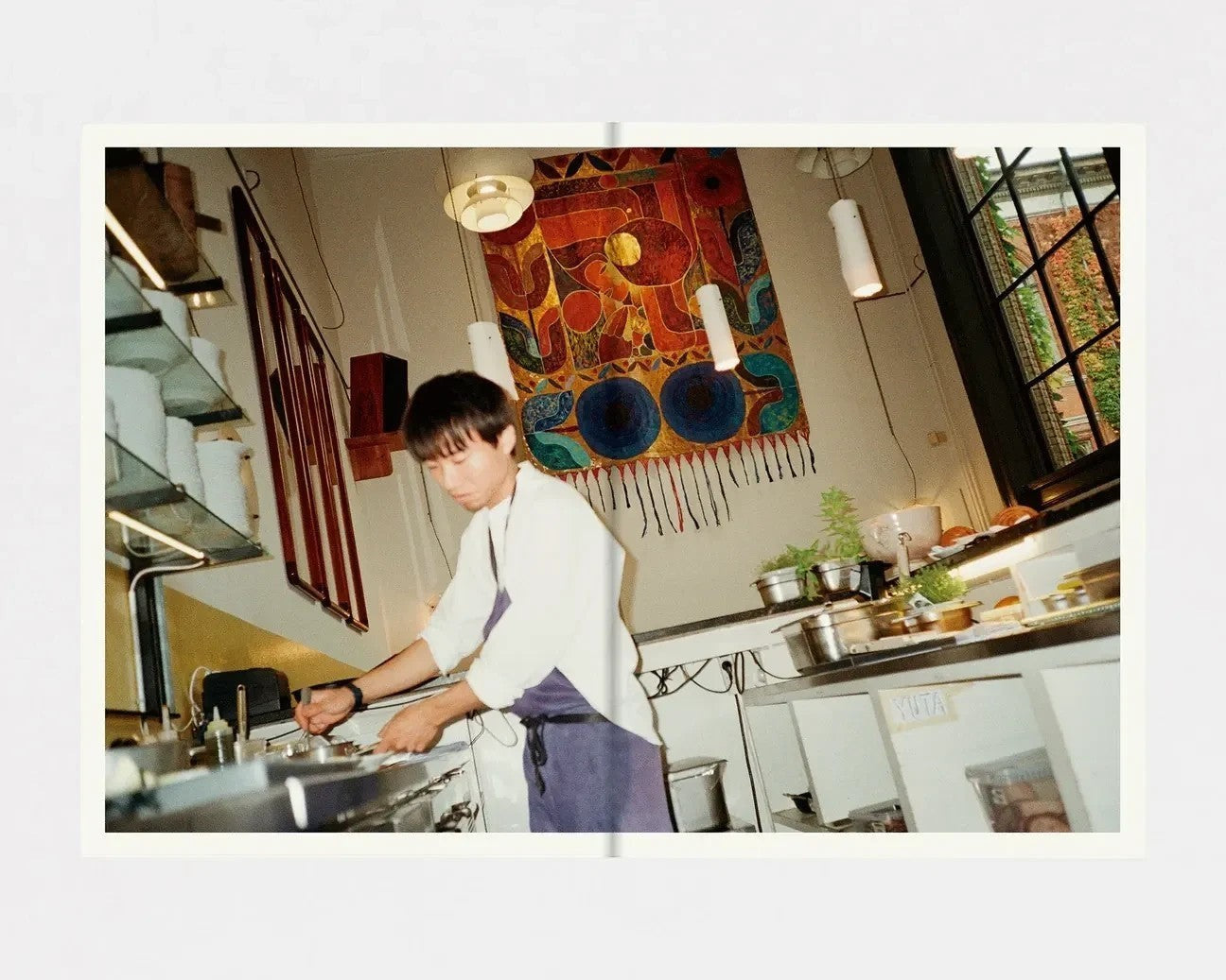 Interior spread of keep-collections Apollo cookbook showing a chef in a kitchen with a vibrant wall tapestry